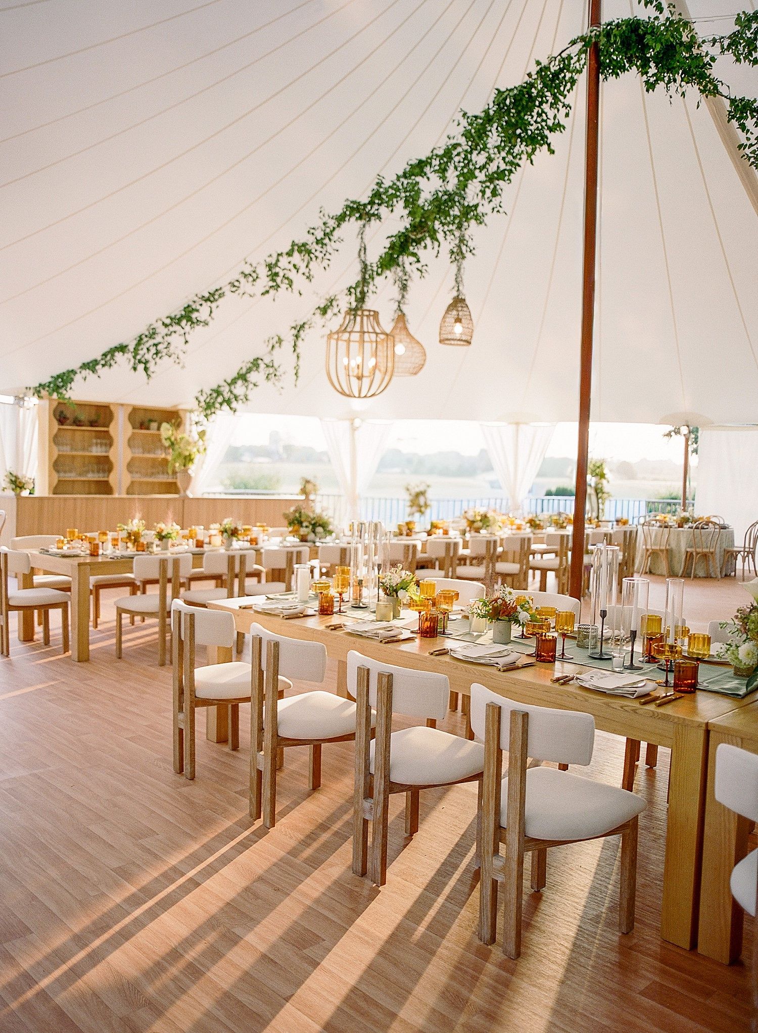 Modern wedding reception space with wooden tables, white chairs, and hanging lights under a spacious white tent.