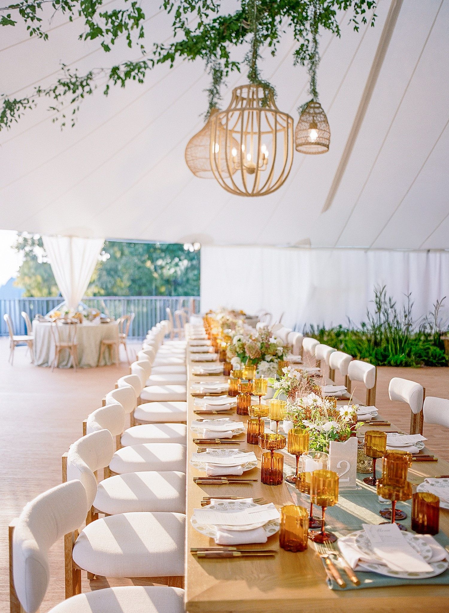 Elegant wedding reception table settings with white linens, amber glassware and natural greenery under pendant lights.