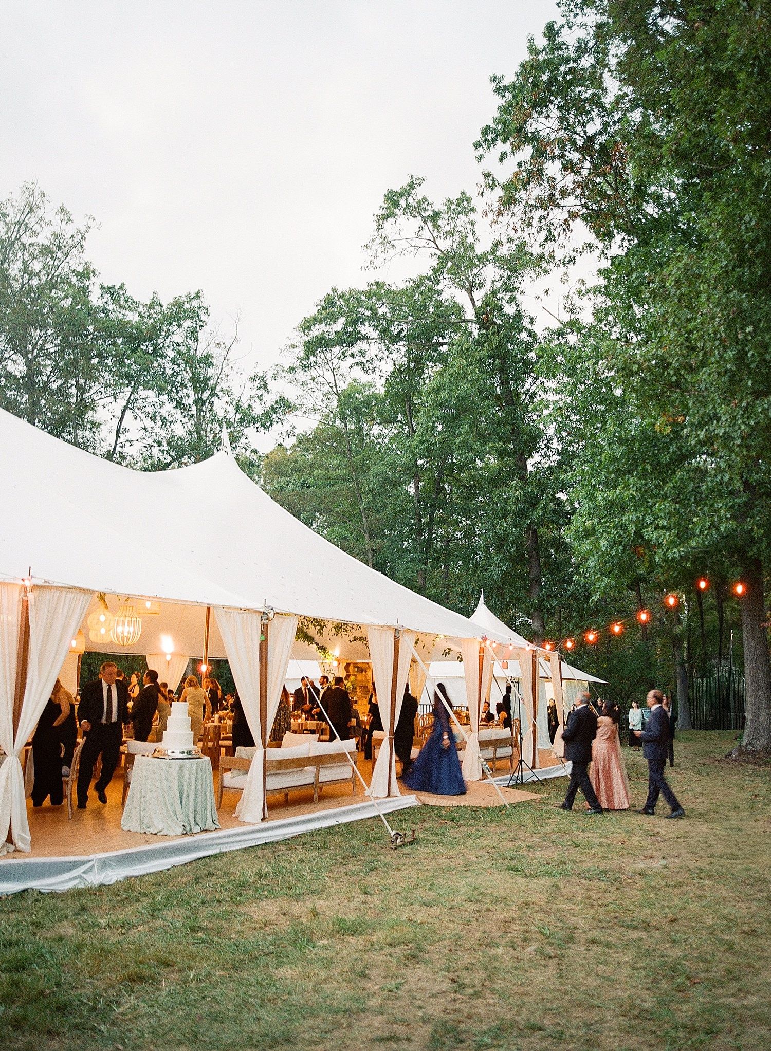 A row of elegant white wedding tents illuminated by string lights at dusk creates a romantic outdoor reception setting.