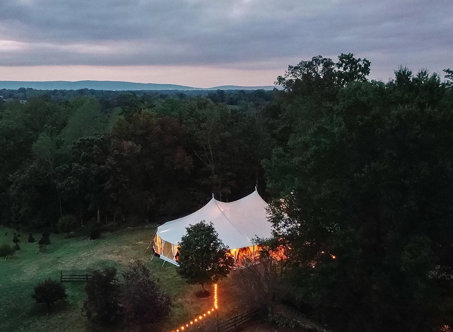 A series of connected white wedding tents illuminated at dusk with string lights creating a magical outdoor setting.