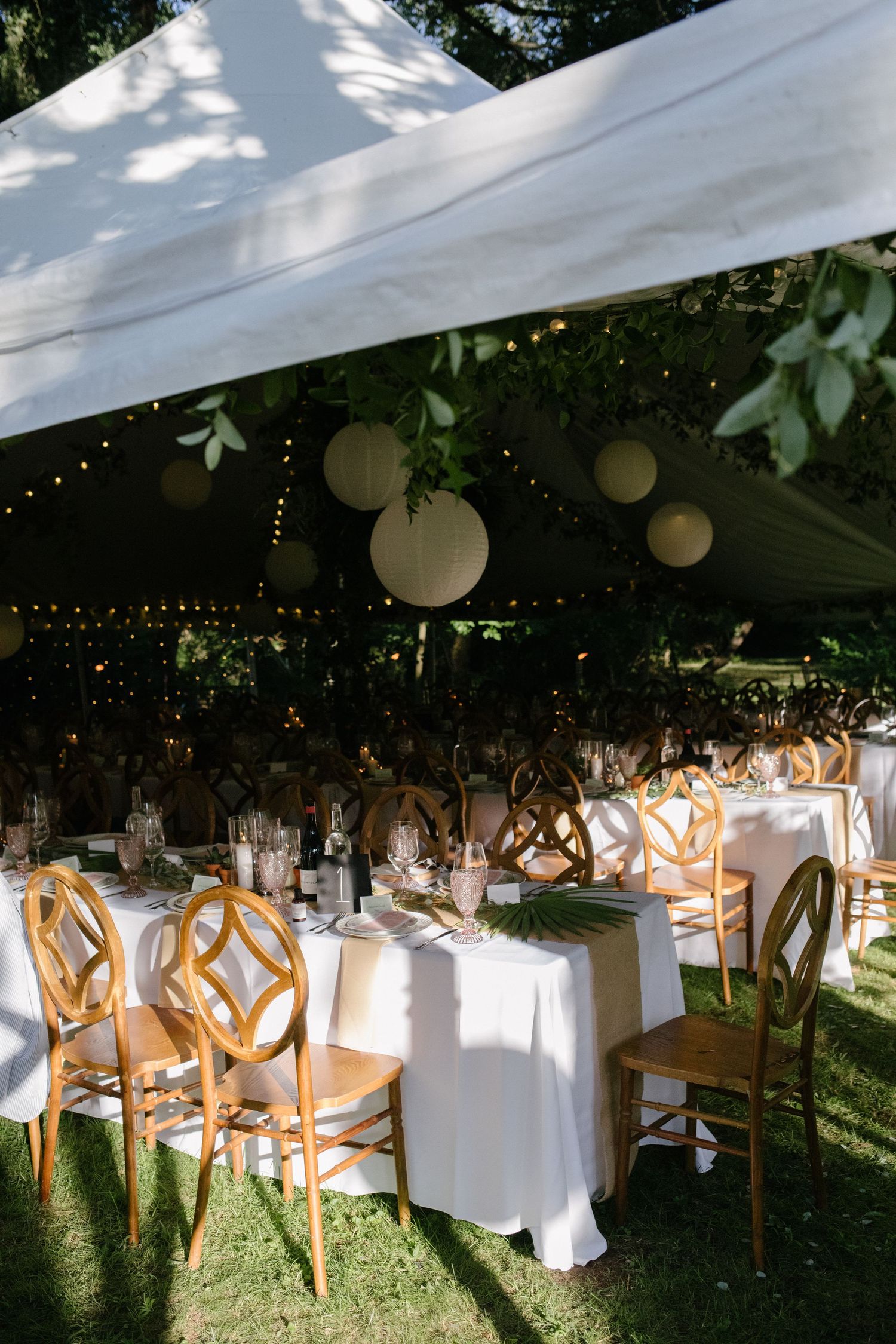 Elegant outdoor tented wedding reception with wooden chairs and white paper lanterns.