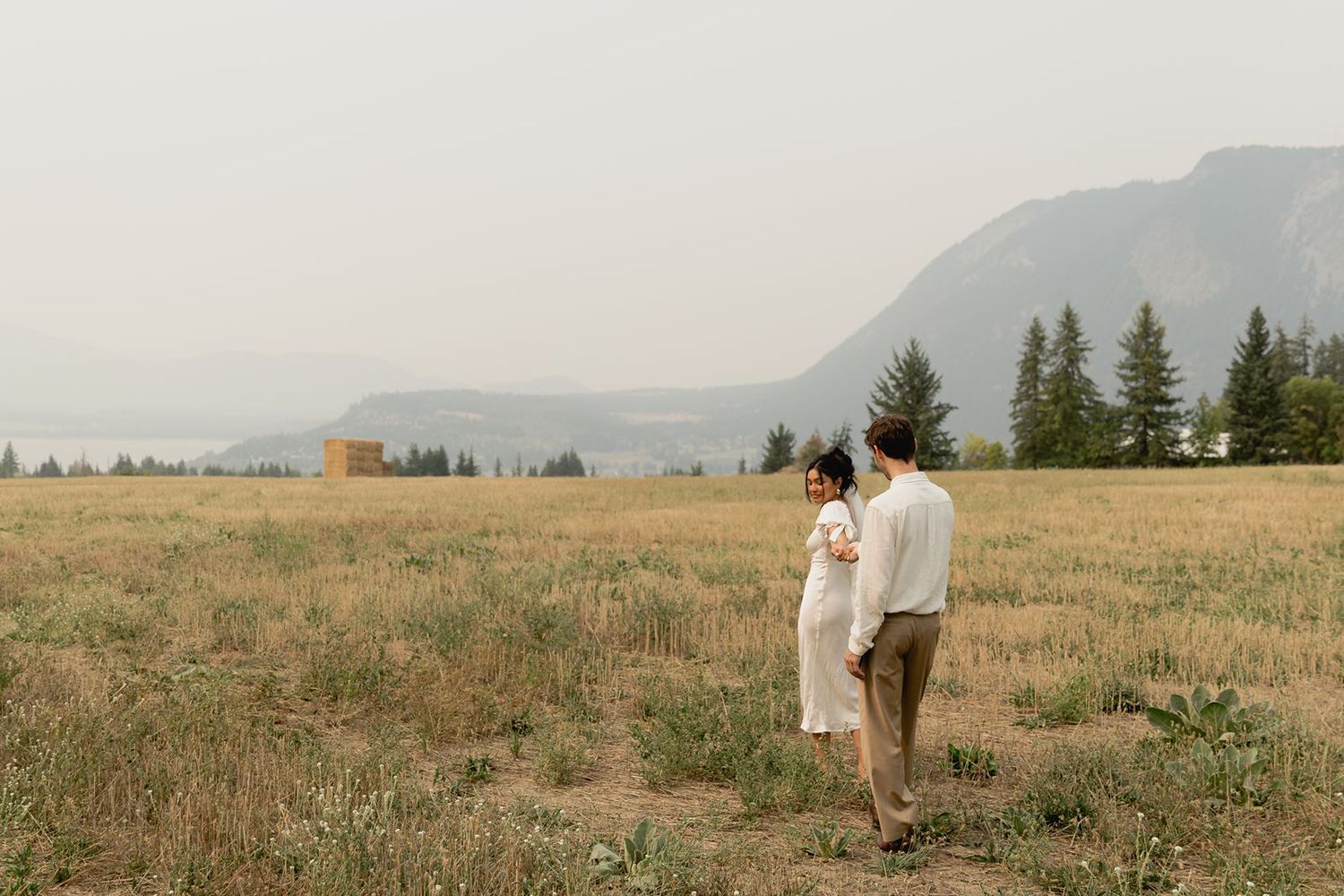 Bride and groom walk hand in hand through golden field with mountain and lake views behind them.