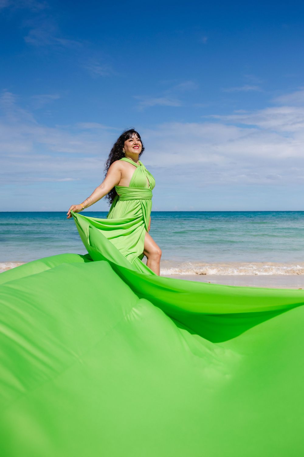 A flowing lime green dress billows dramatically against a pristine beach landscape with turquoise waters and blue sky.