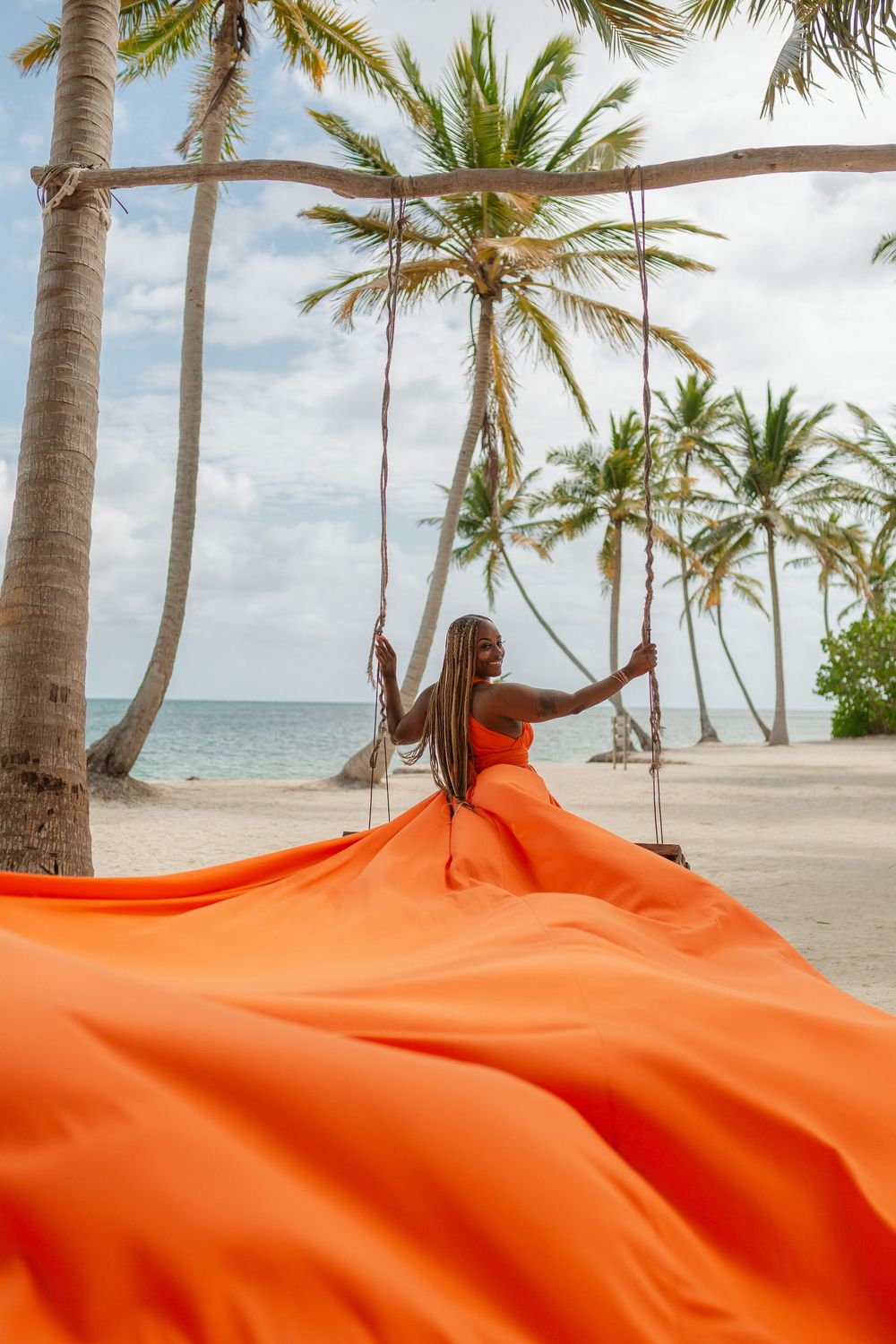 Bright orange fabric billows in the breeze against a tropical beach backdrop with palm trees and turquoise ocean waters.