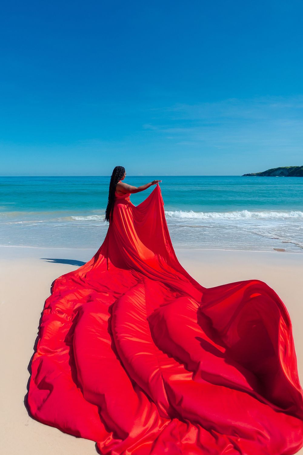 Red flowing fabric billows against pristine white sand beach and turquoise ocean waters under bright blue sky.