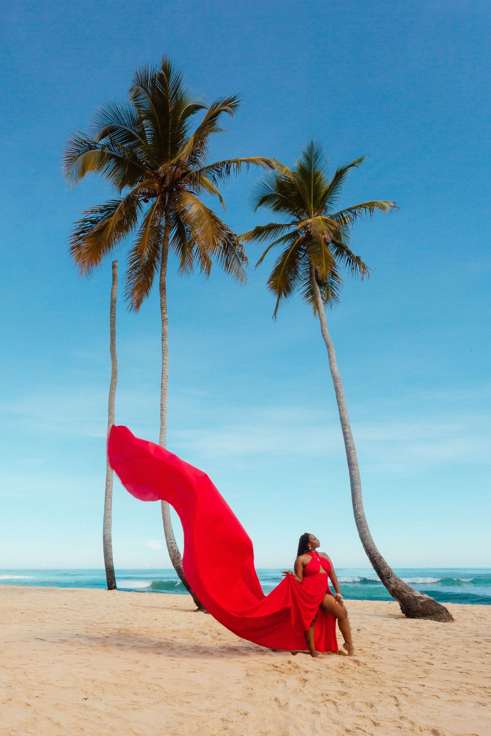 Red flowing fabric creates dramatic movement against palm trees and white sand beach on a bright sunny day.