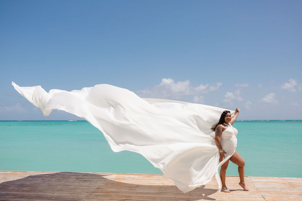 A flowing white wedding dress billows in the ocean breeze against turquoise Caribbean waters and blue sky.