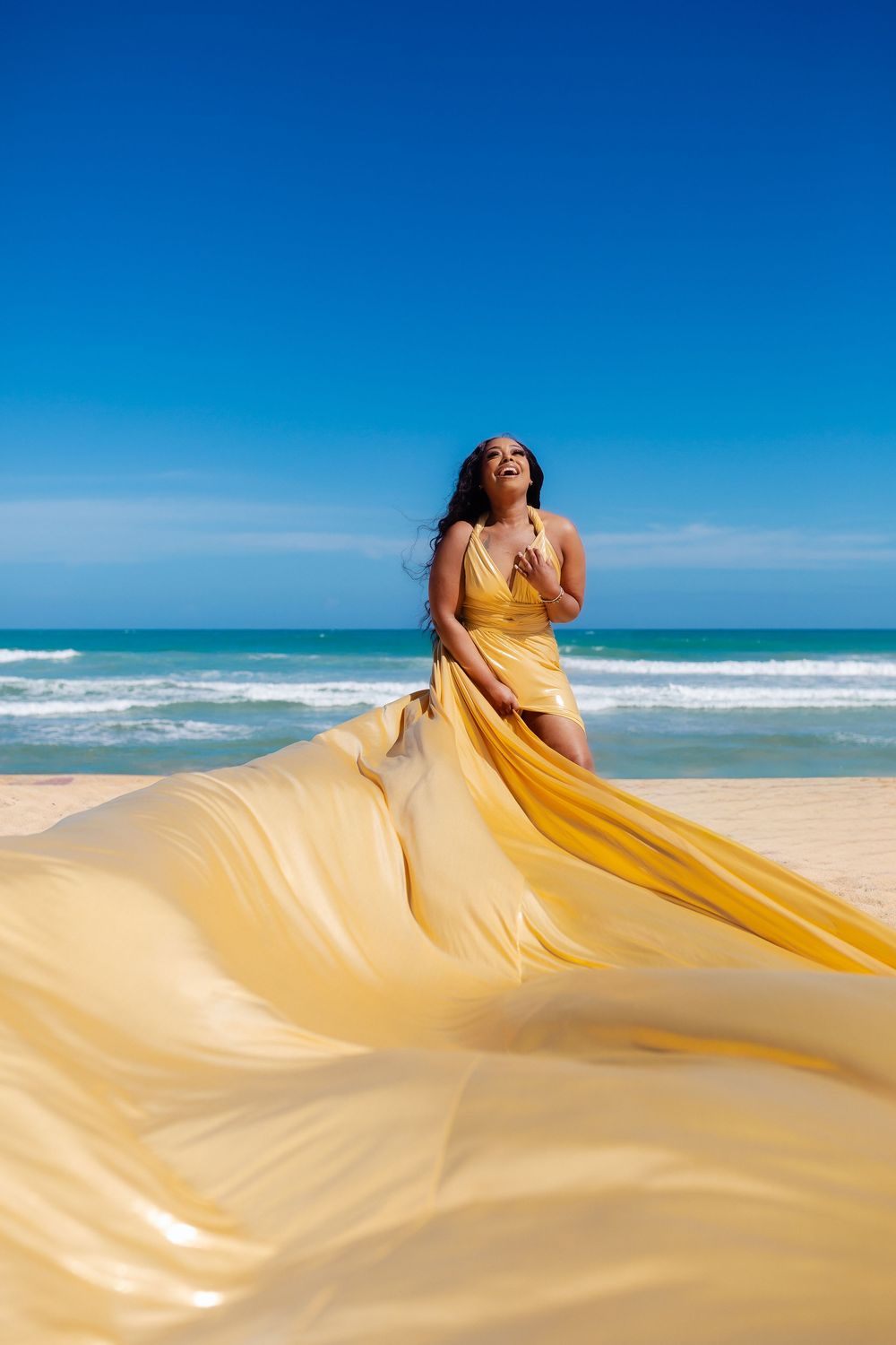 A flowing gold dress billows dramatically on a sunny beach as waves crash in the background against bright blue skies.
