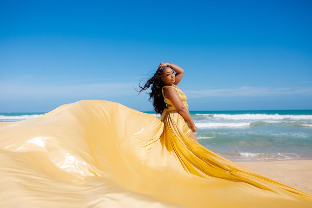 Gold flowing fabric billows gracefully against a bright blue sky and sandy beach backdrop.