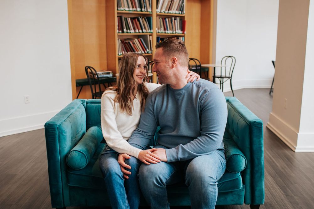 Couple sits close together on a teal velvet couch in a library setting.