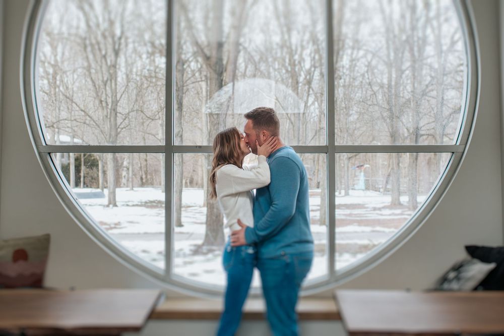 A person in a teal sweater sits by a large circular window overlooking a snowy winter landscape.