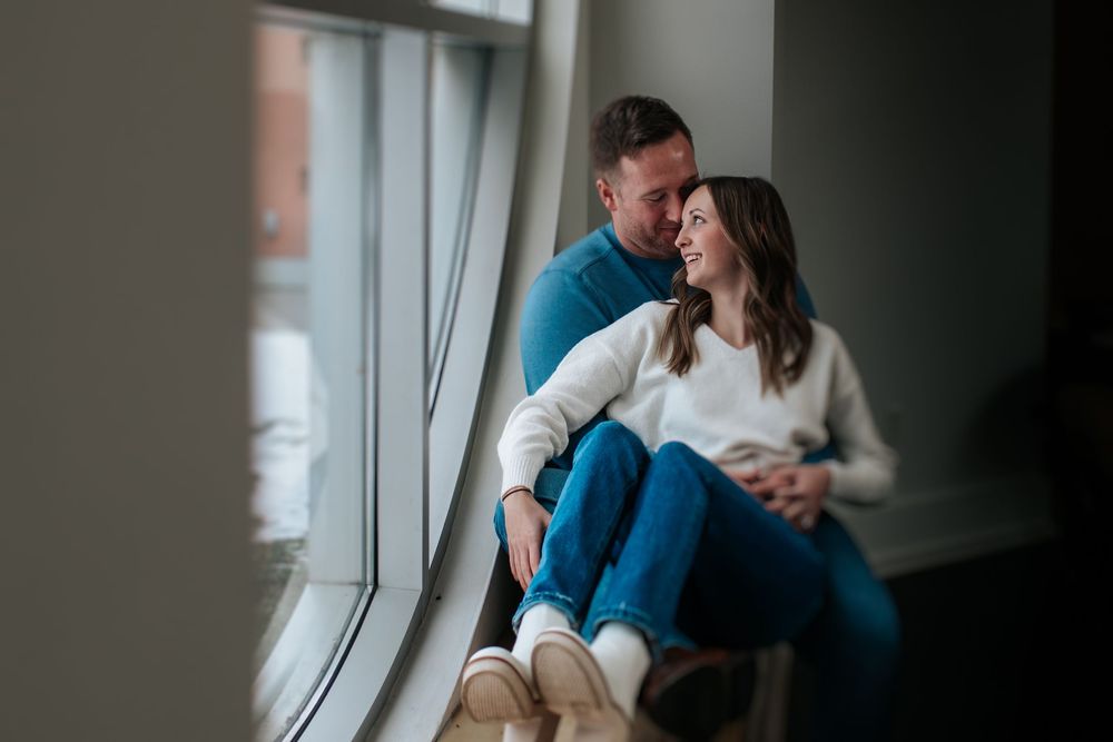 A couple sits alone by a window in a dimly lit room, wearing blue jeans and looking contemplative.