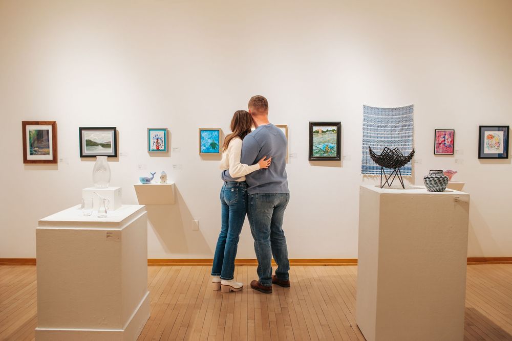 A couple view framed artwork displayed along a wall with pedestals featuring sculptures in the exhibition space.