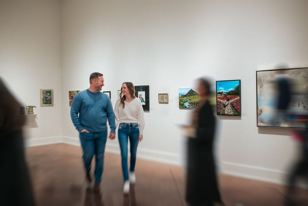Couple walks through an art gallery with white walls displaying framed artwork.