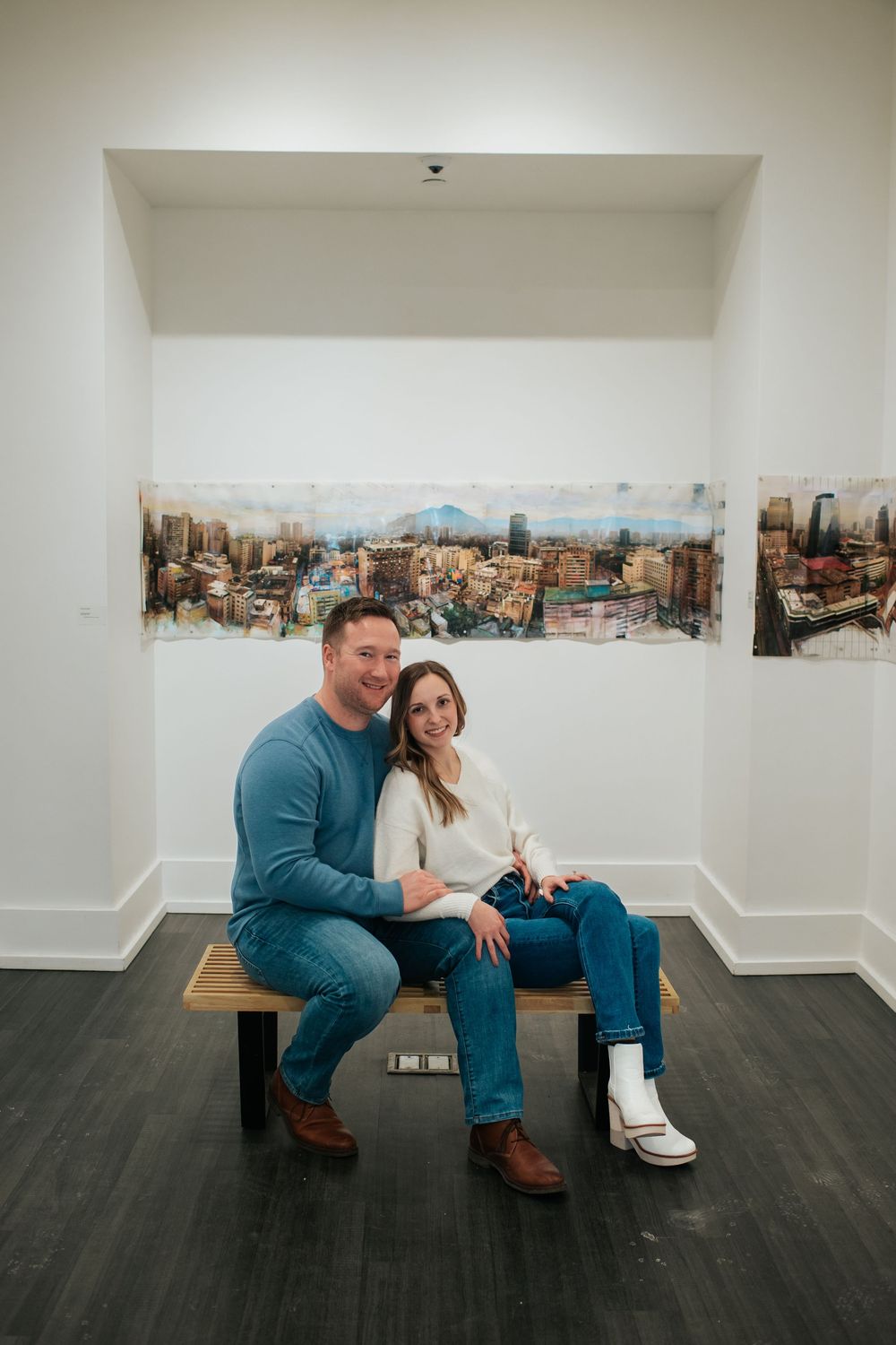 Couple poses casually in an art gallery with a panoramic cityscape painting behind them.