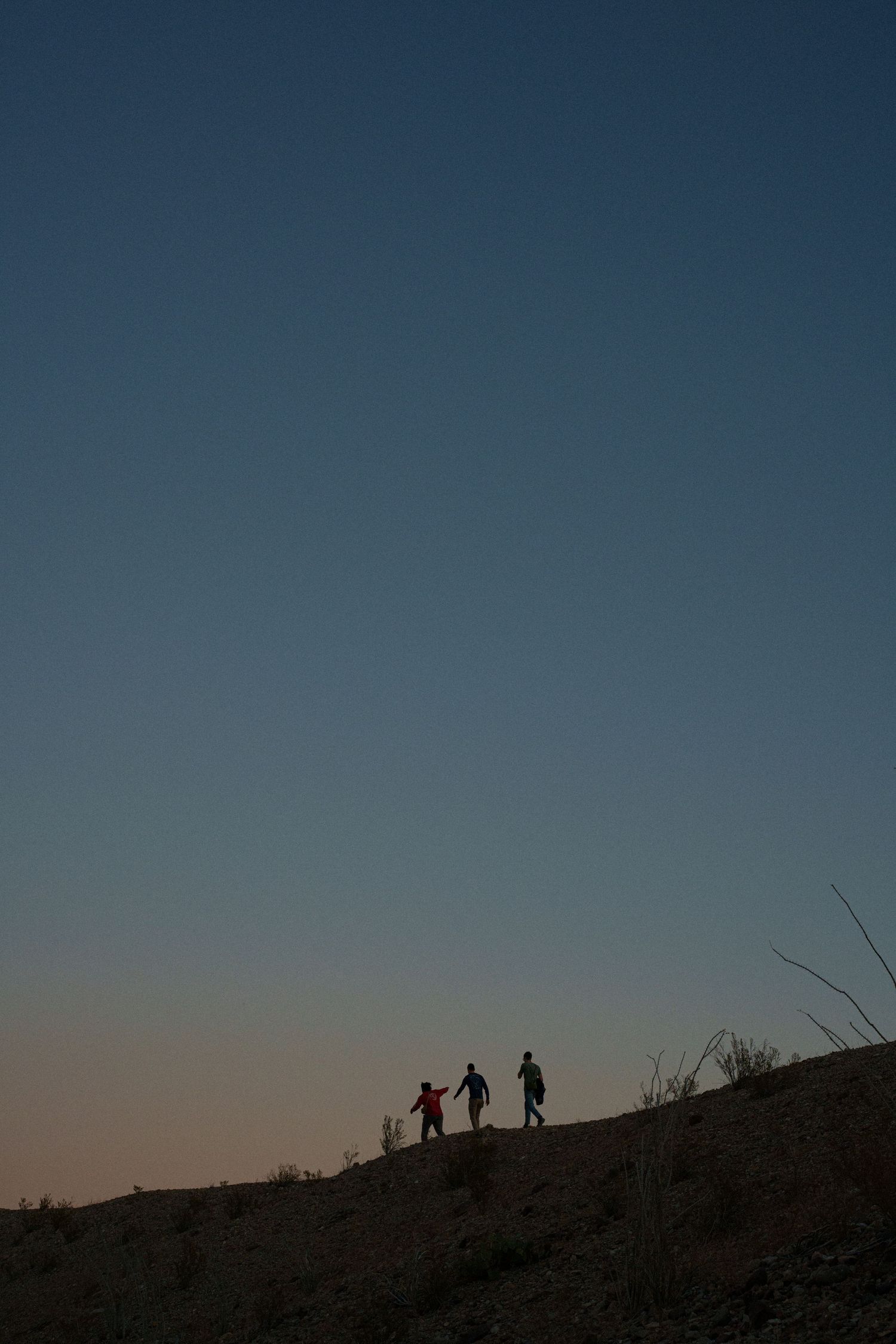 Three silhouetted figures stand on a hilltop against a dusky blue sky at sunset.