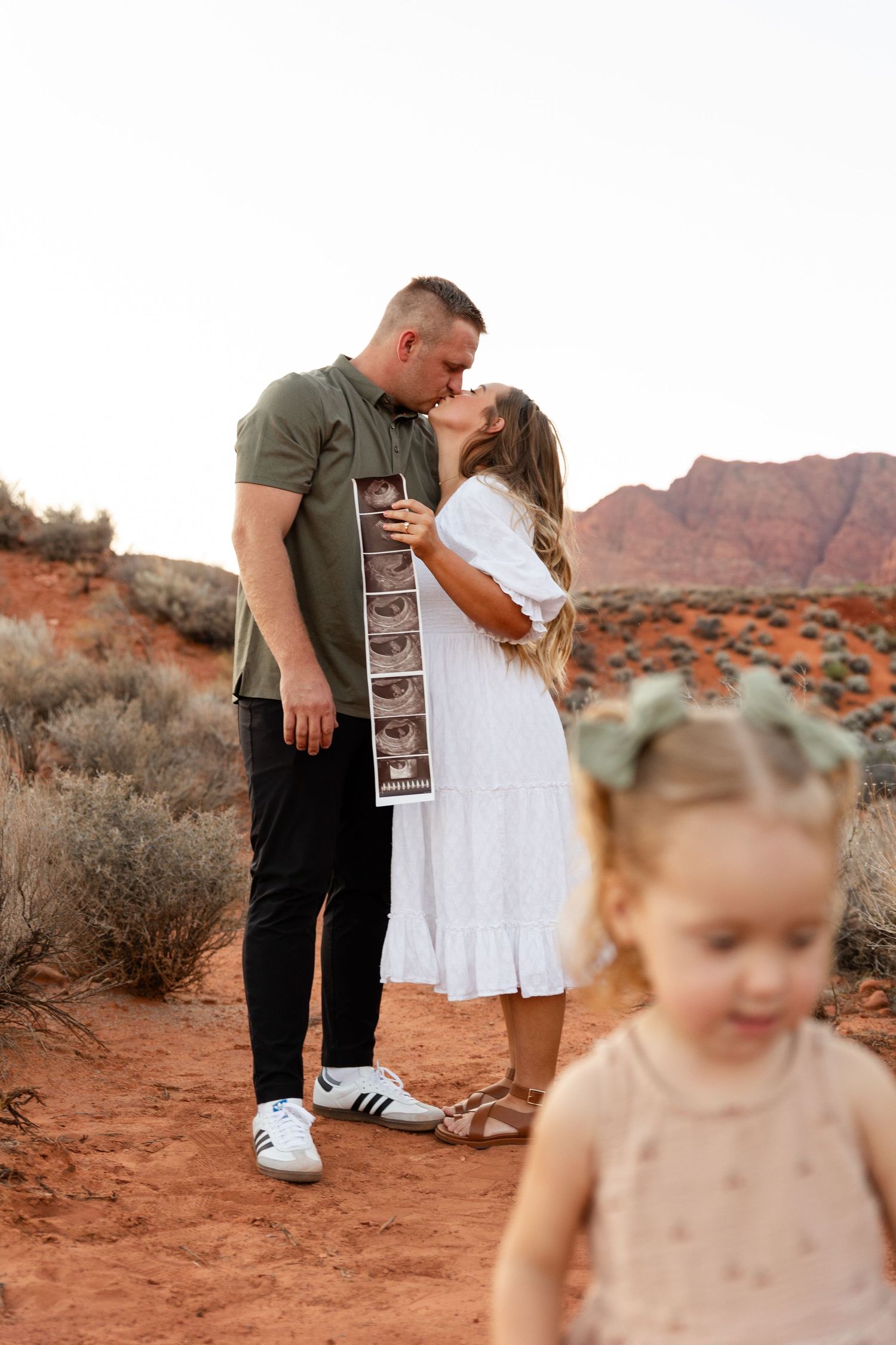 mom and dad holding sonogram kissing in the background. focus on them. 2 year old girl walking away in the foreground out of focus