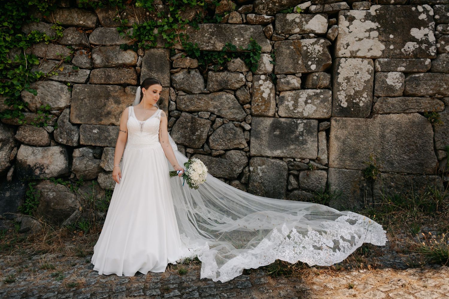 A bride in a flowing white wedding gown poses against a rustic stone wall in a series of romantic bridal portraits.
