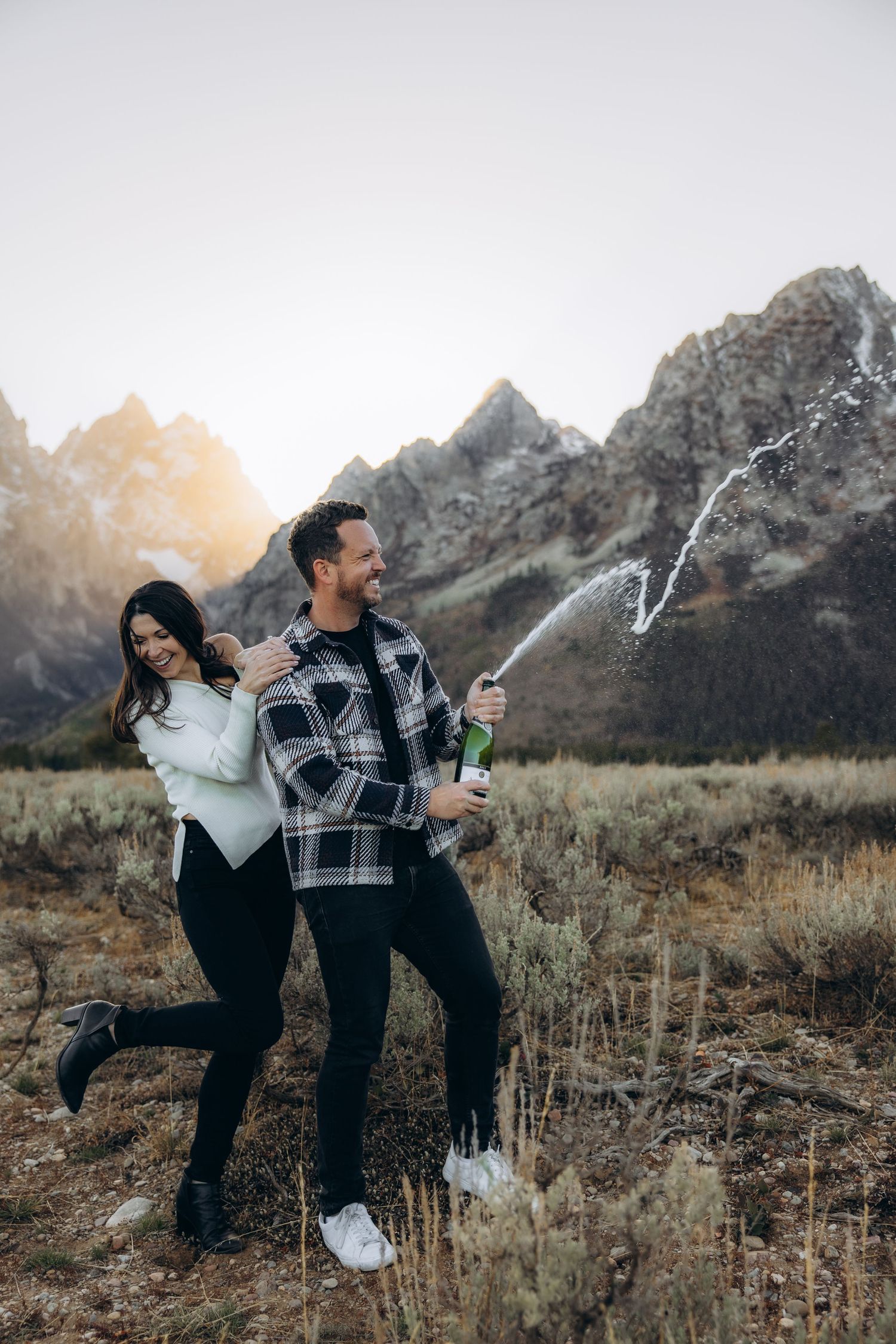 A couple sprays champagne together in a field with mountains in the background during sunset.