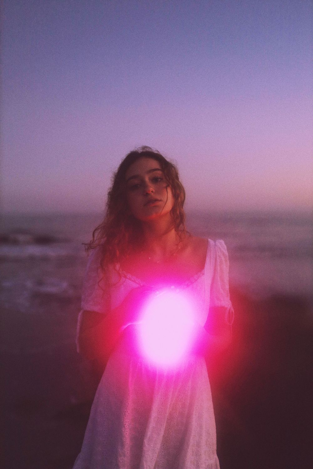 A dreamy beach scene at dusk with a glowing pink orb of light illuminating a figure in a white dress.