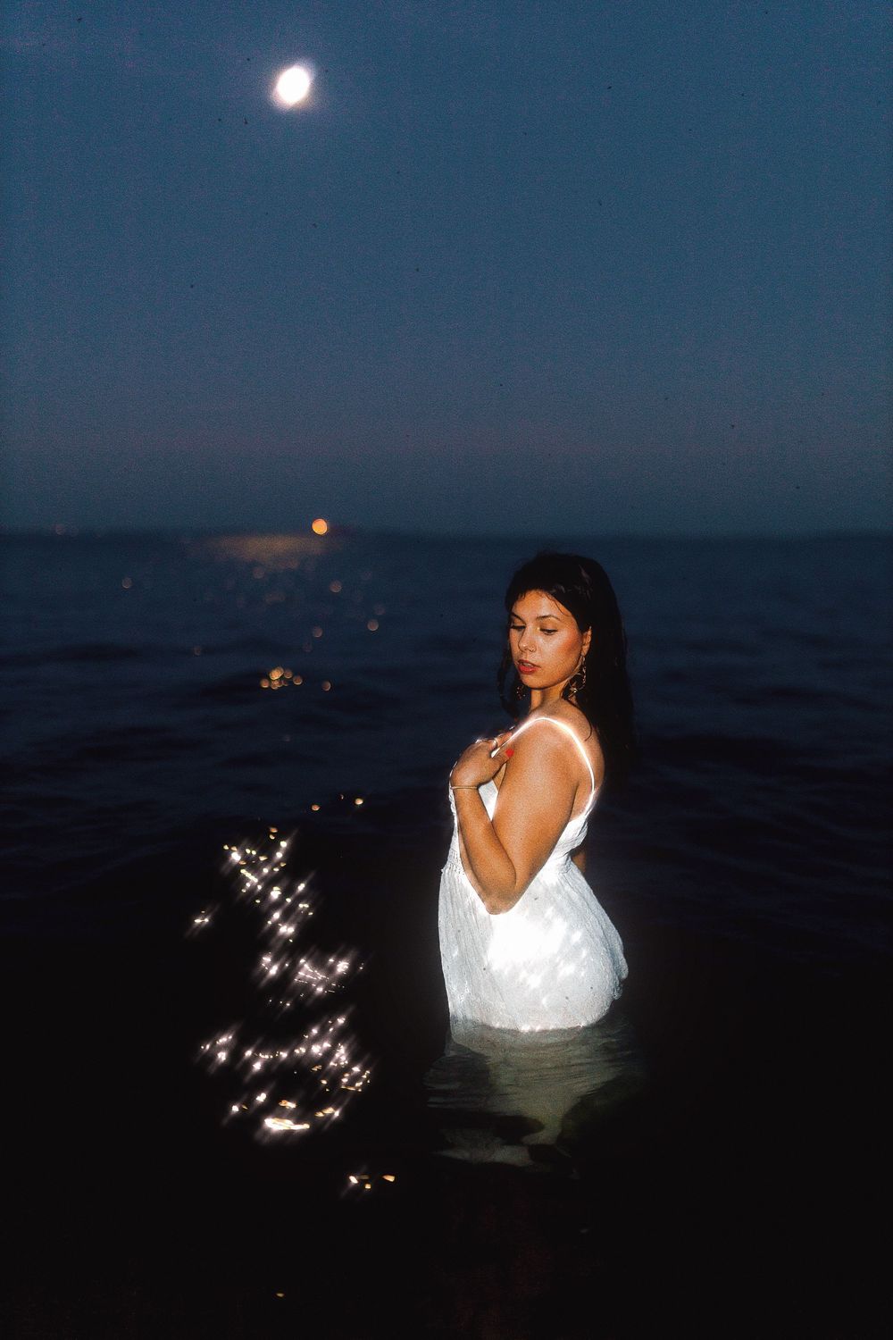 Artistic nighttime photoshoot in water featuring a white dress and sparkling lights under a moonlit sky.