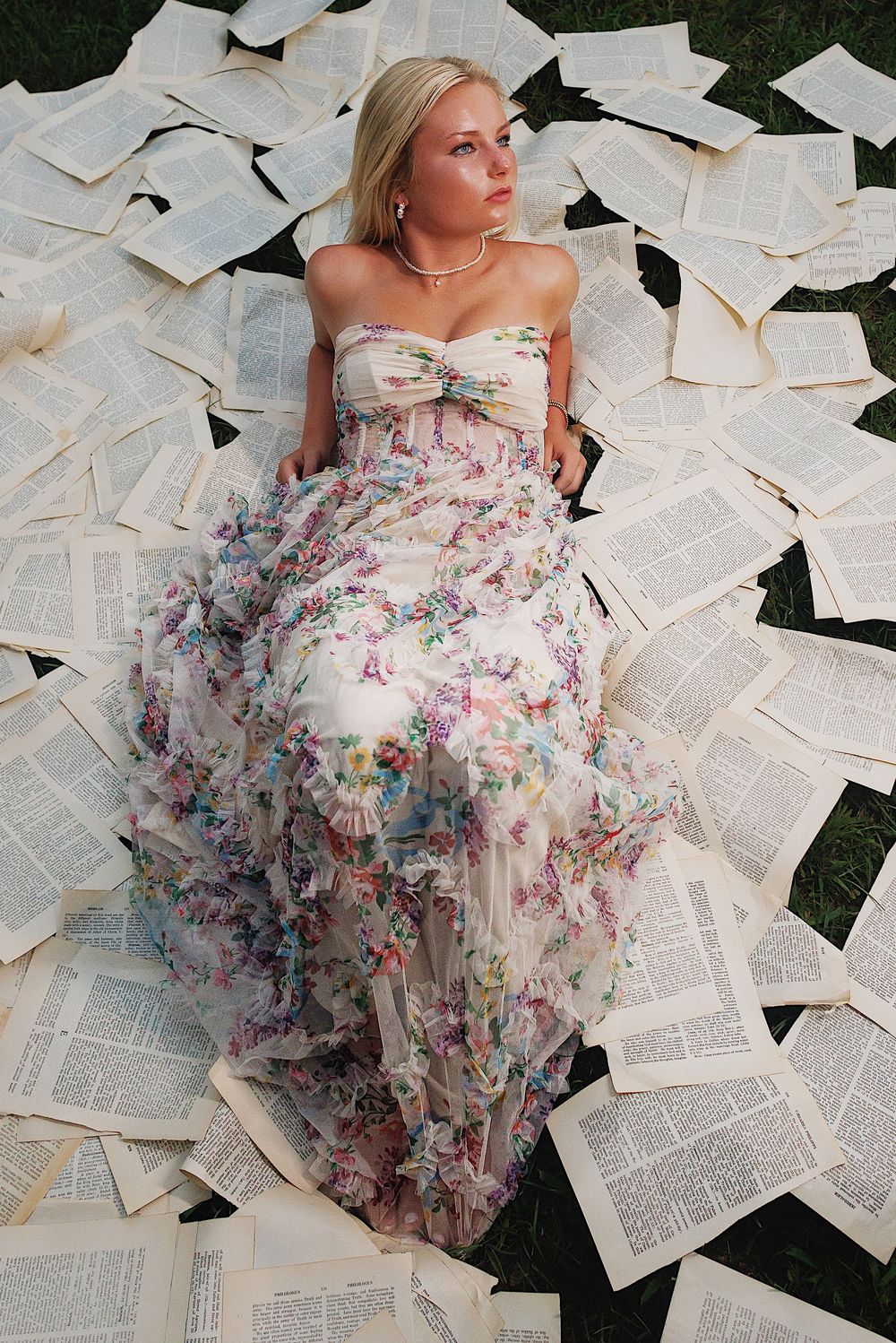 A floral organza dress spreads across scattered book pages on grass in an artistic fashion photoshoot.