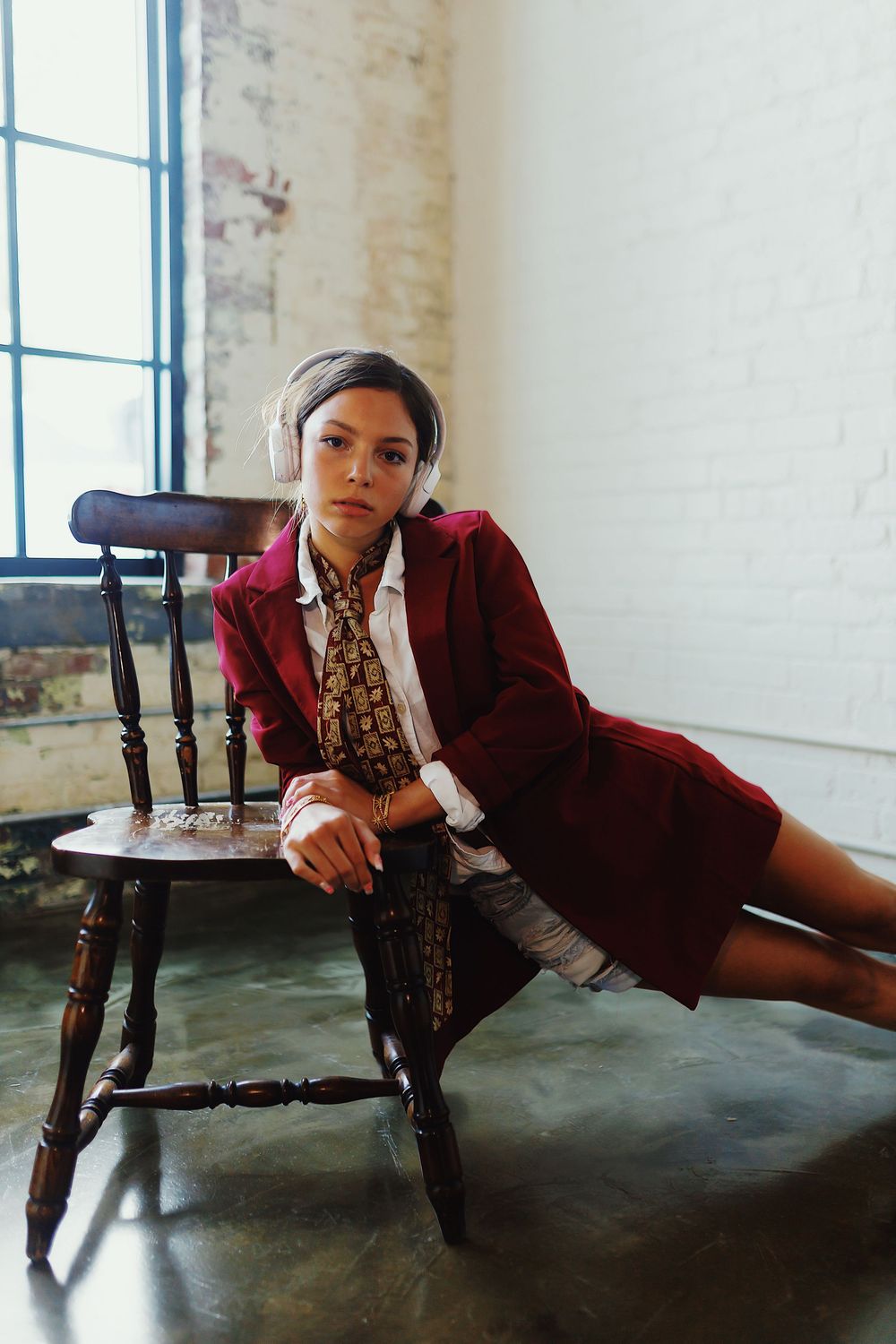 A model in a burgundy coat and patterned dress poses elegantly on a vintage wooden chair against a white wall.