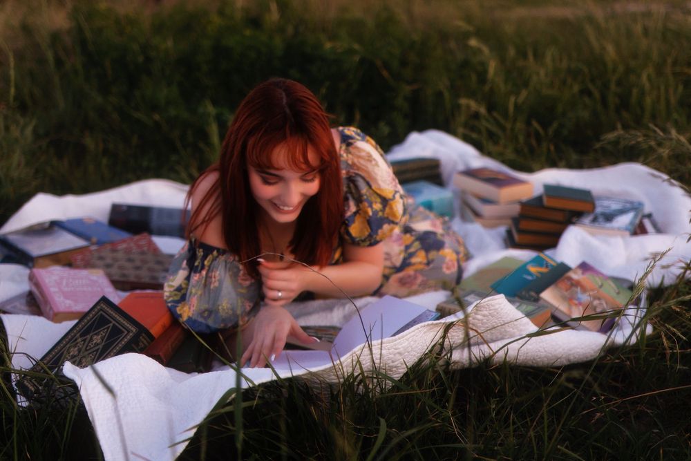 A person lies on a white blanket outdoors during sunset while reading a book in a peaceful setting.