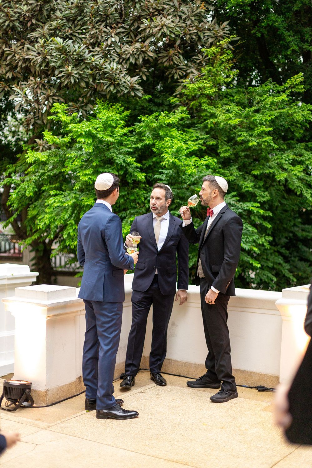 Three men in formal attire having a conversation on an outdoor terrace.