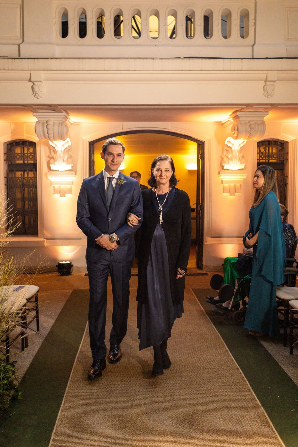 Wedding party members walk down a carpeted aisle in an elegant indoor venue with ornate architectural details.