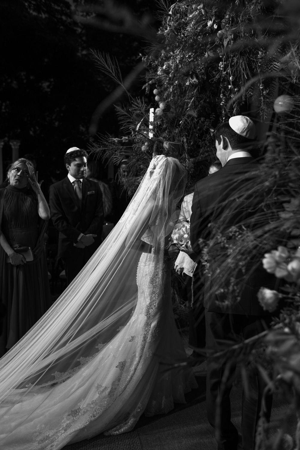 A black and white photo of a wedding ceremony with a bride in a long flowing veil surrounded by greenery.