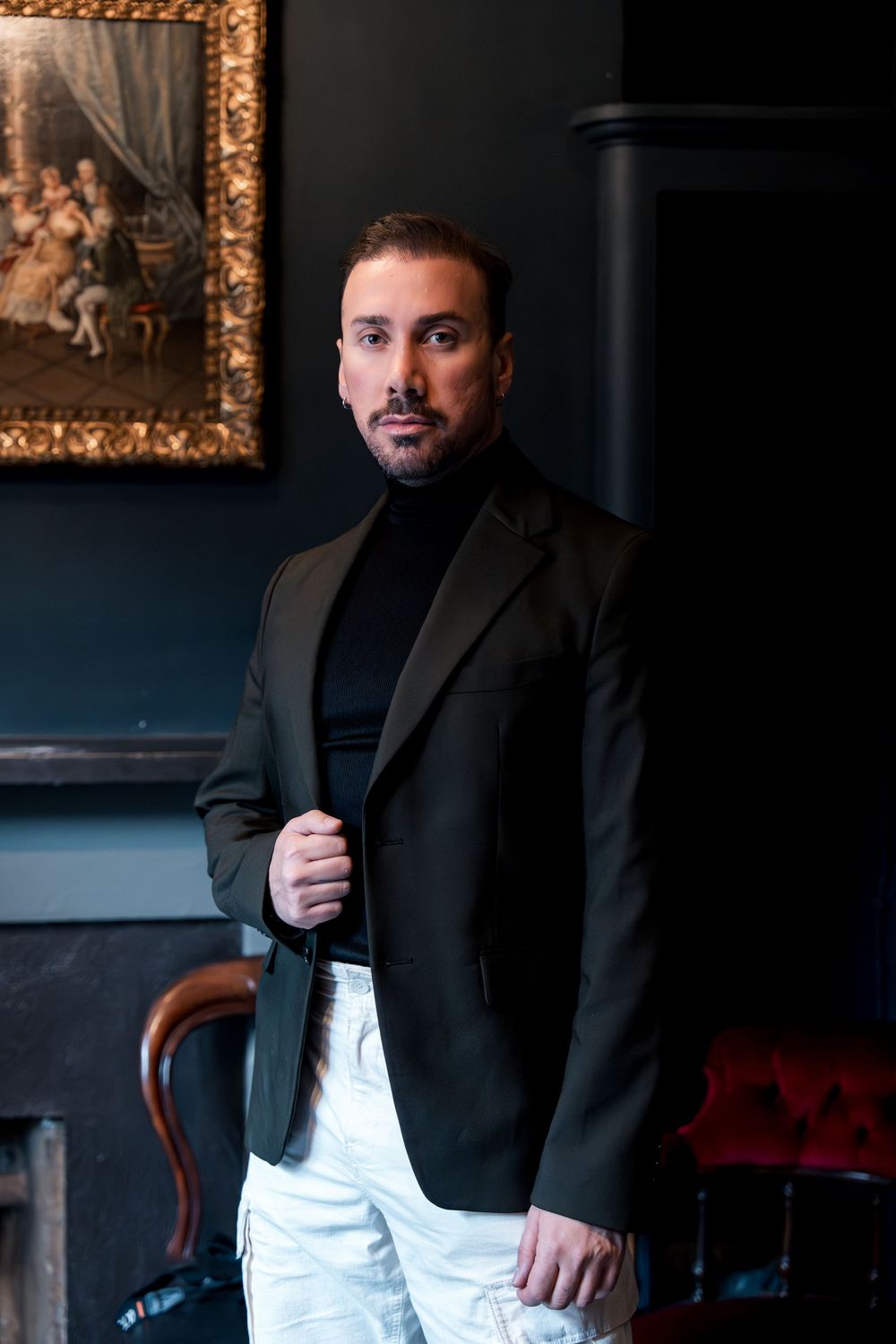 Stylish portrait in a dark room with navy walls featuring a black blazer, turtleneck and white pants.