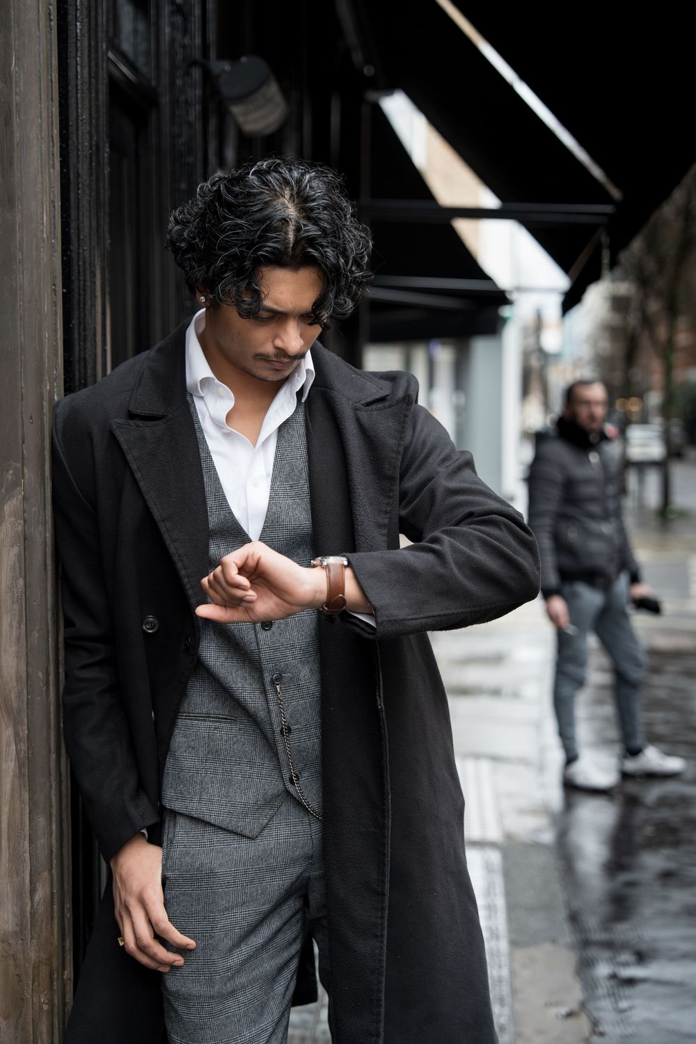 A person in a black coat and gray vest poses against a wall in a series of stylish fashion photographs.