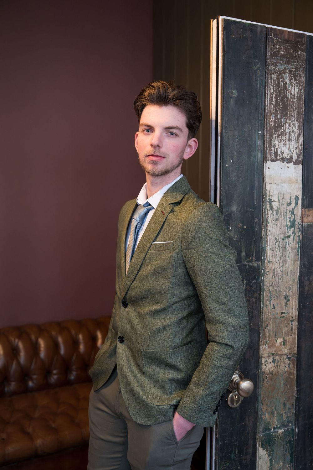 A person in a tailored olive green suit poses against a rustic wooden pillar in a dimly lit room with leather seating.