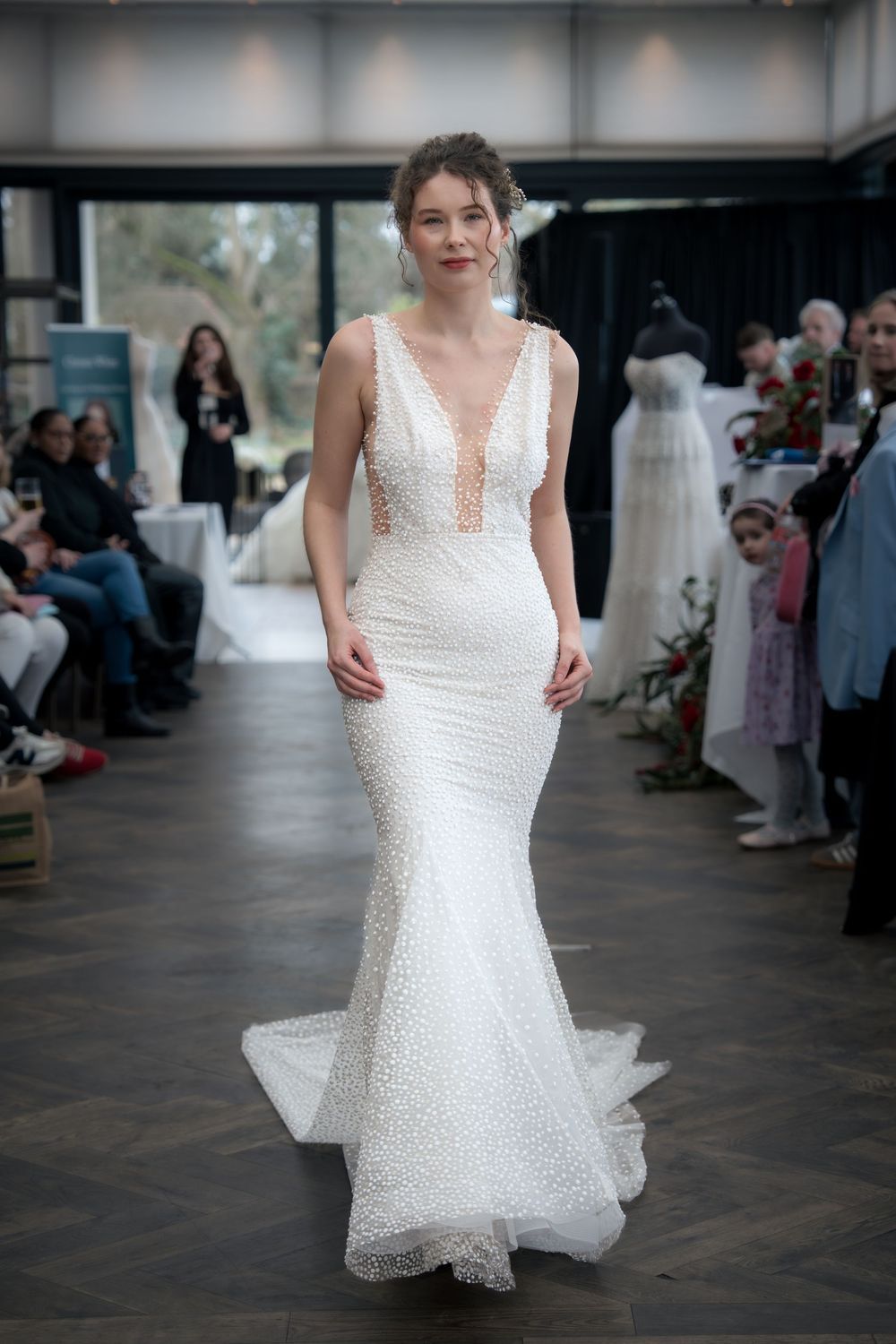 A model walks the runway in a fitted white sequined wedding gown with plunging neckline at a bridal fashion show.