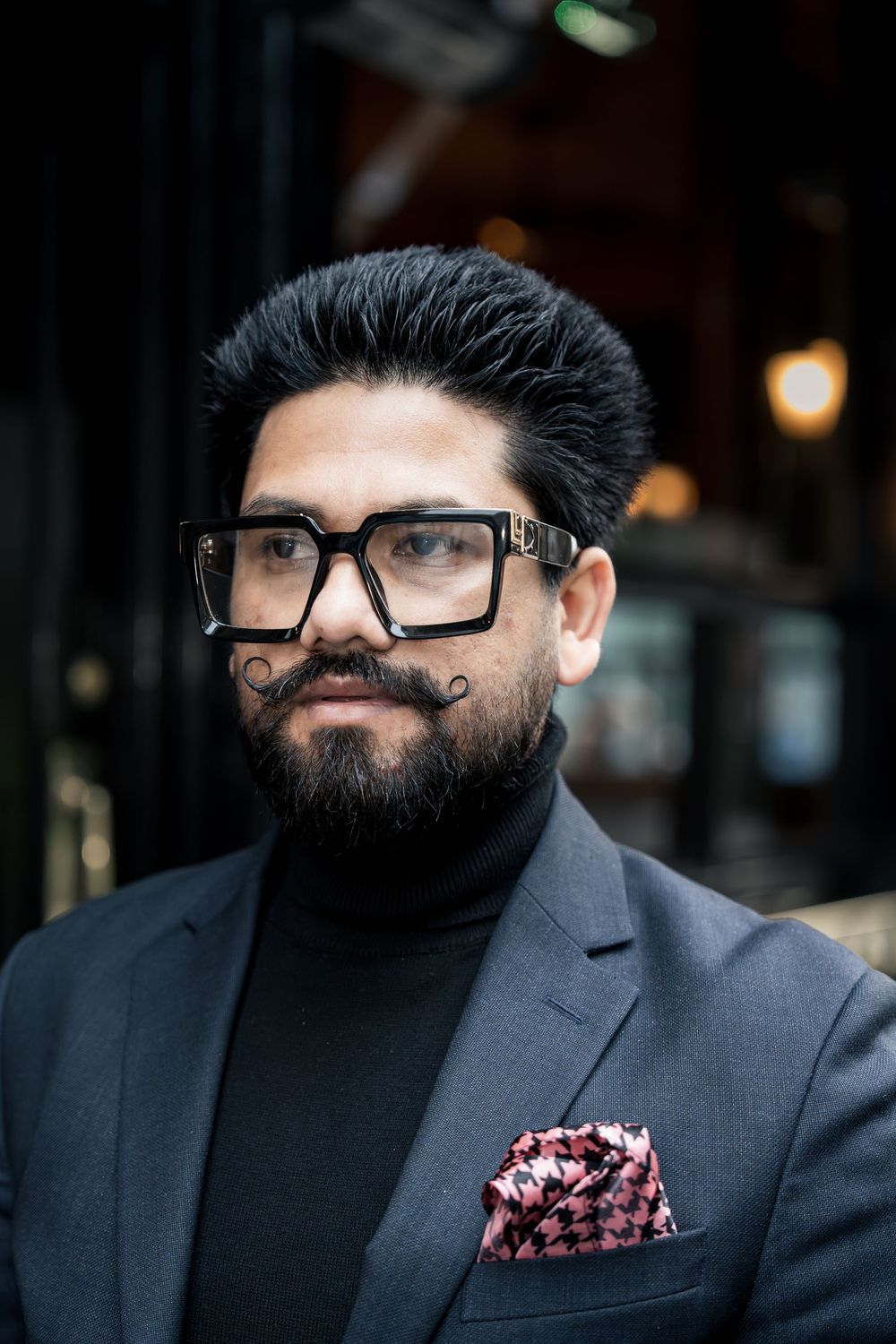 A bearded person wearing black-rimmed glasses and a gray suit jacket with a patterned pocket square looks to the side.