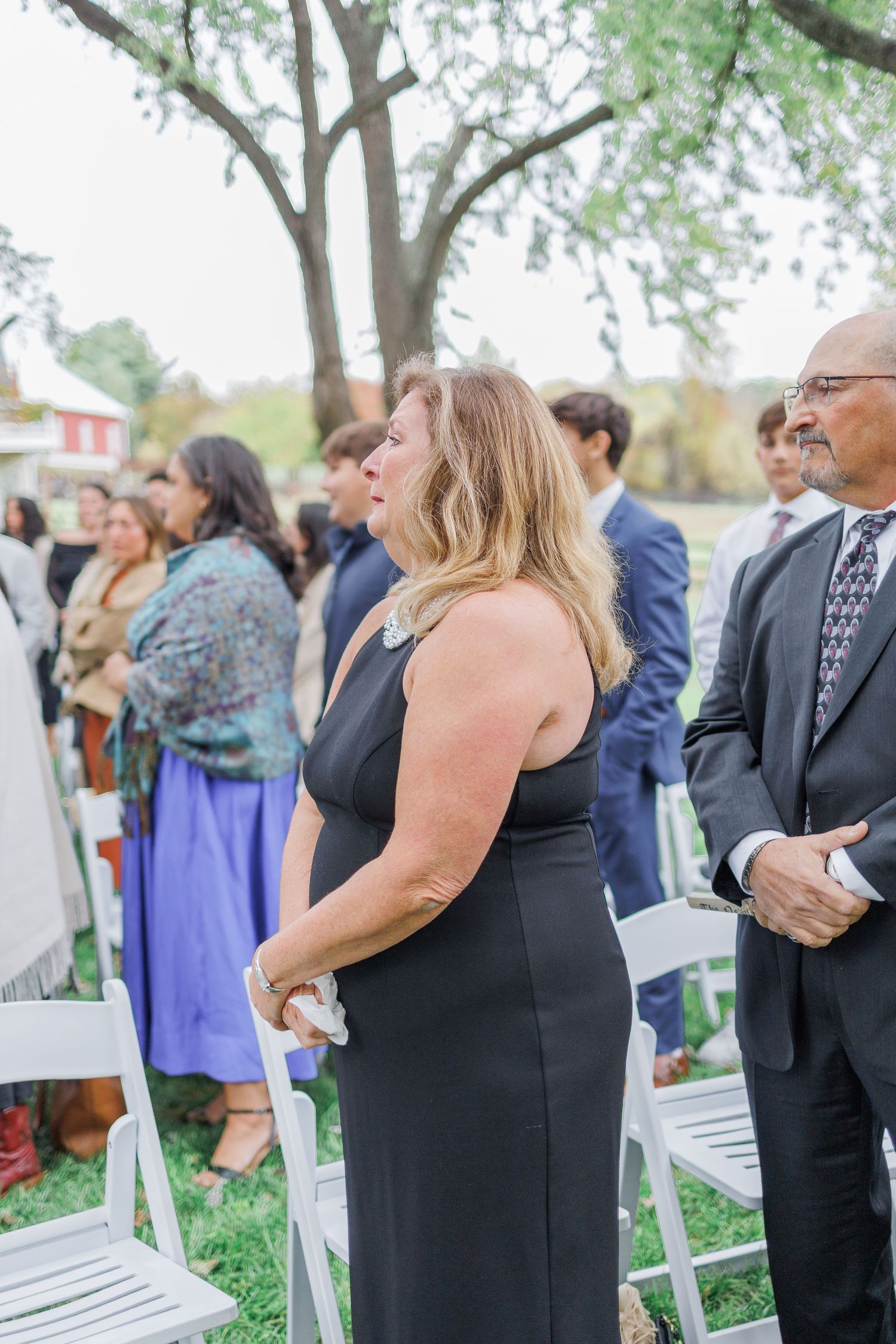 Wedding guests gather outdoors on a lawn with white chairs during a ceremony on a sunny day.