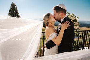 A romantic wedding couple embraces on a scenic balcony with flowing white veil and mountain views in the background.