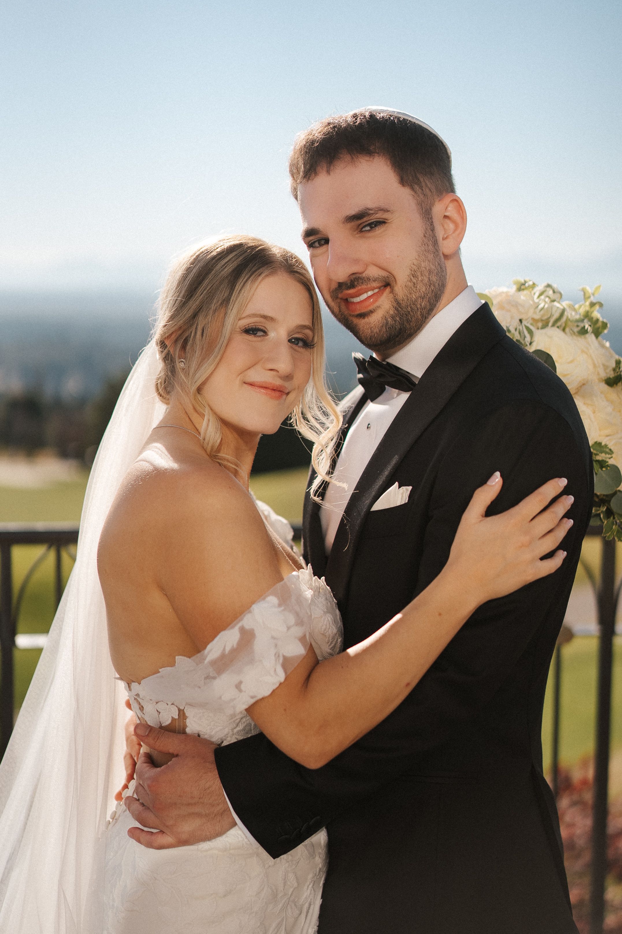 A newlywed couple embraces outdoors on their wedding day in elegant attire against a scenic mountain backdrop.