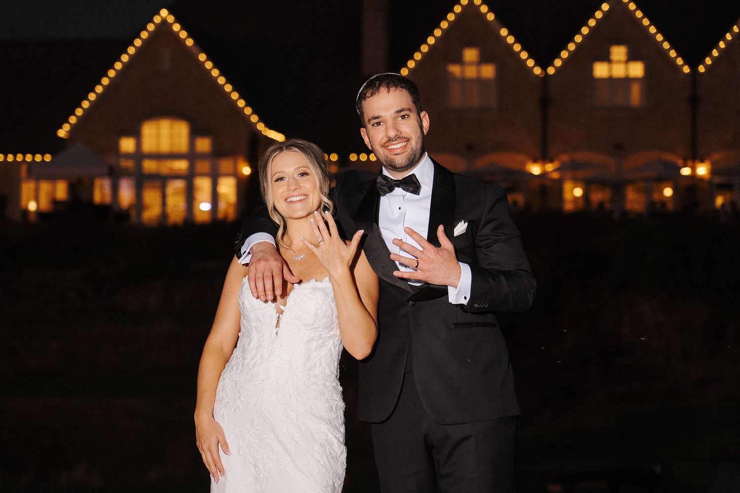 Newlyweds pose together at night in front of illuminated venue with twinkling lights in background.