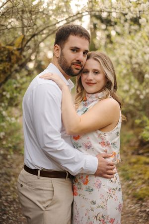 Romantic couple embraces on a wooded path surrounded by blooming spring trees and dappled sunlight.