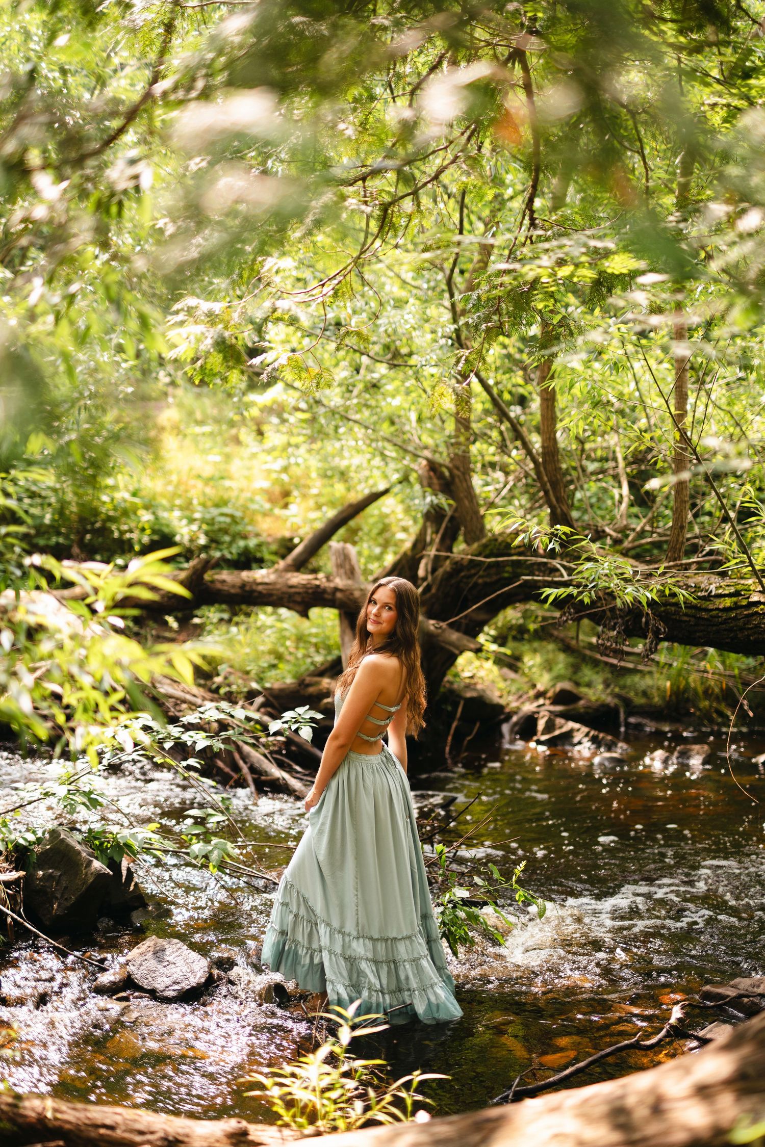 Smiling blonde in emerald green tank top poses outdoors against colorful summer foliage backdrop.