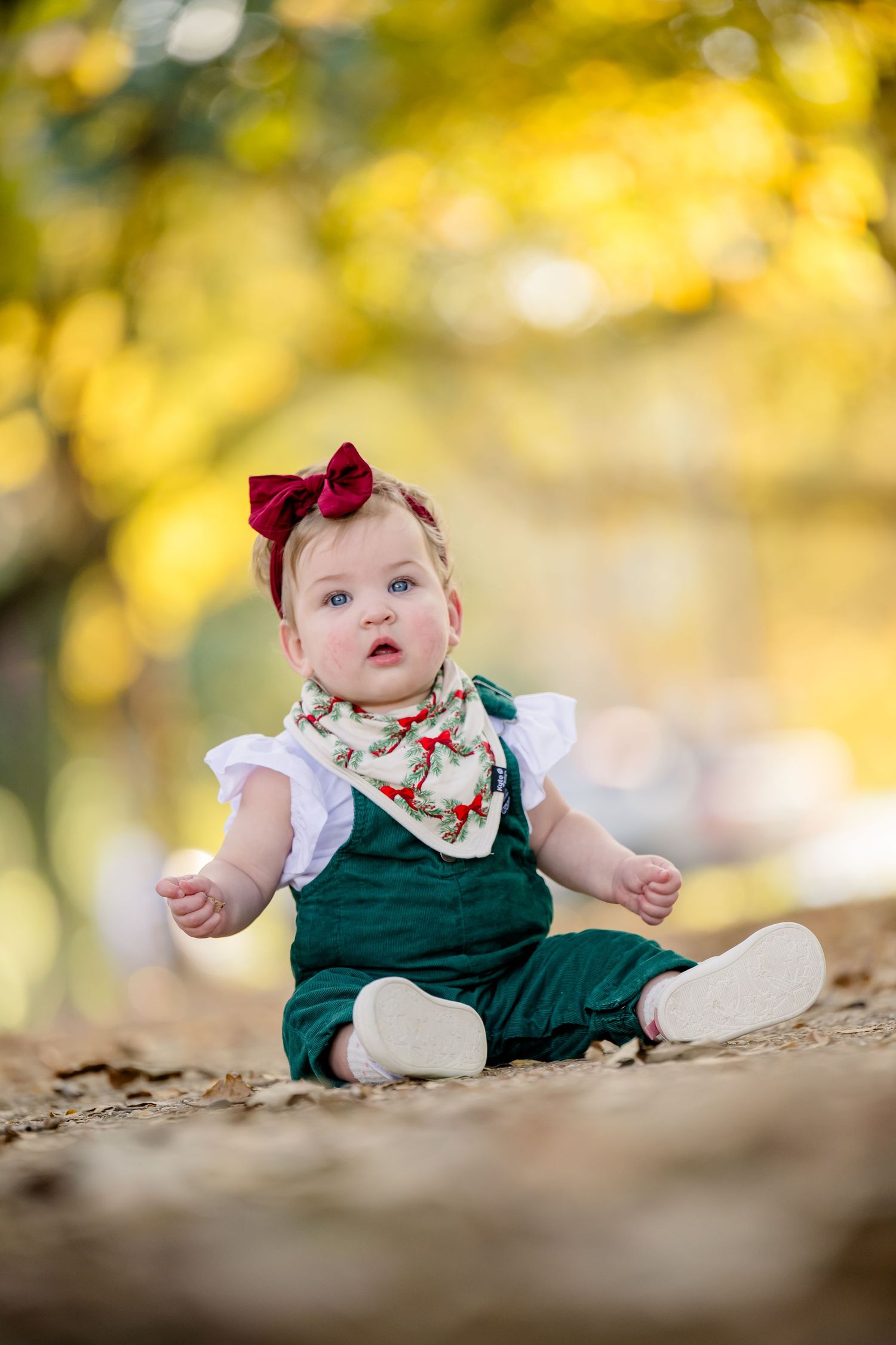 Baby in green dress with red bow sits in autumn leaves during outdoor photo session with golden bokeh background.