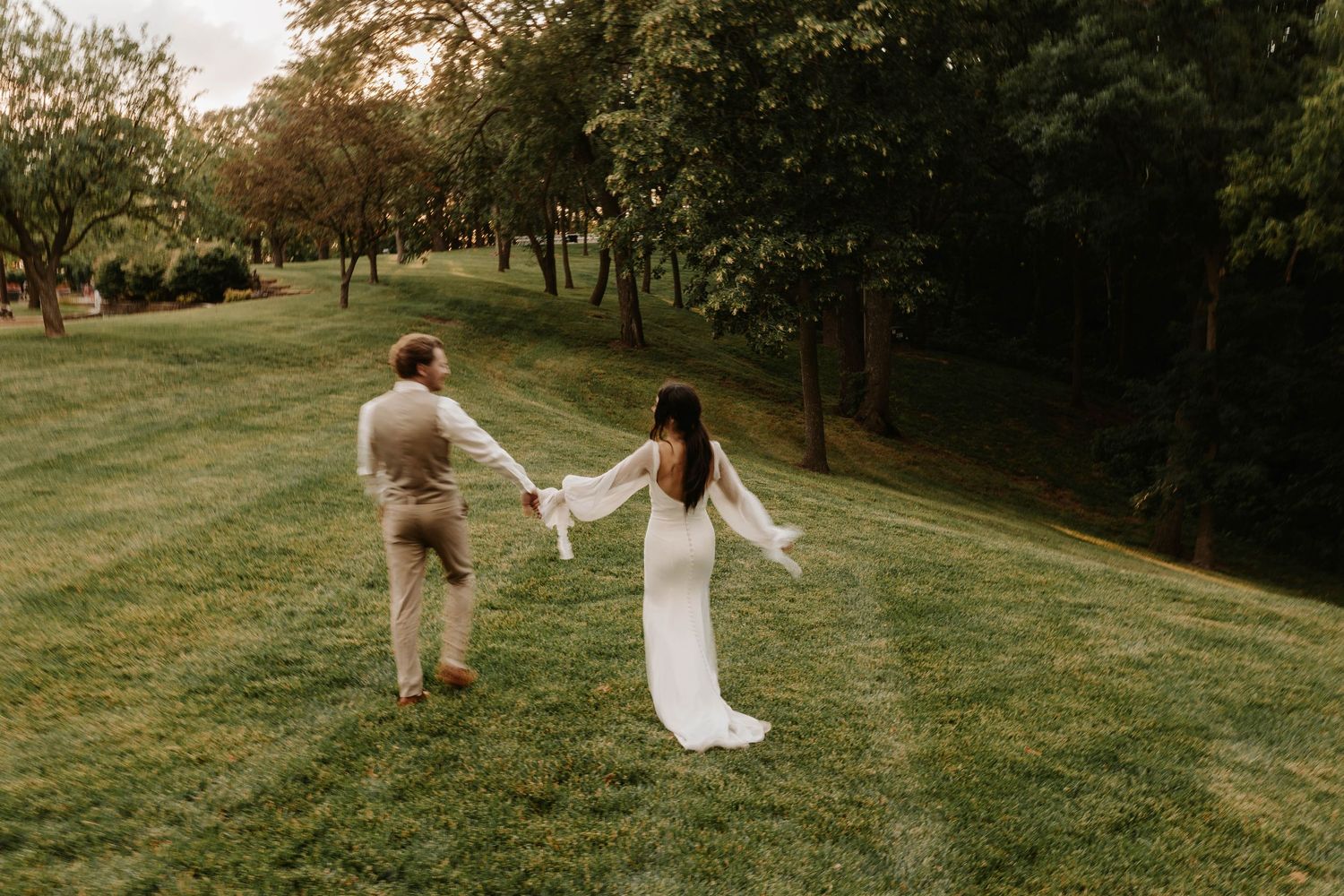 A couple walks hand in hand through a scenic green field during golden hour at sunset.