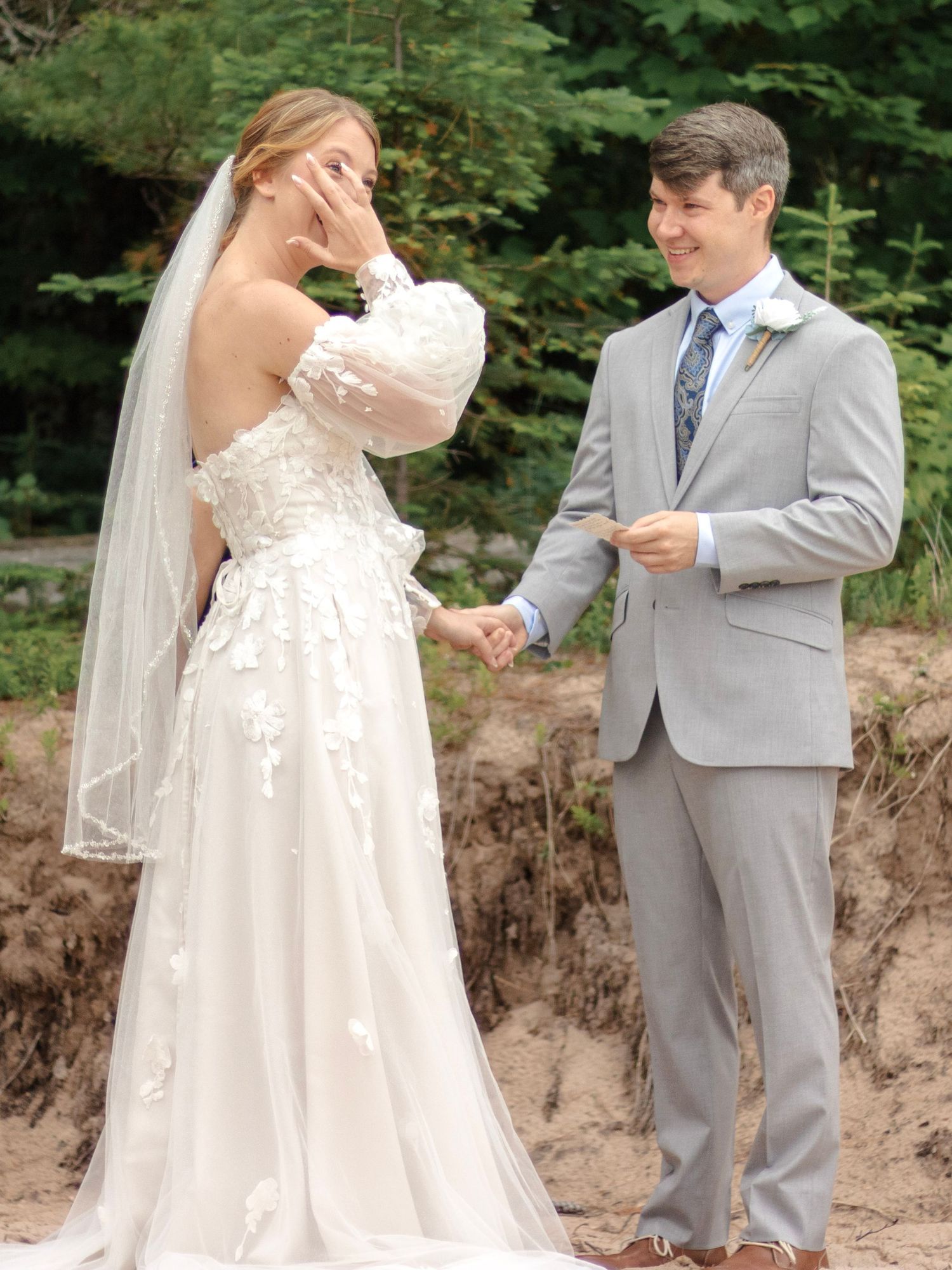 Bride in lace wedding gown and veil holds hands with groom in gray suit during outdoor beach ceremony.