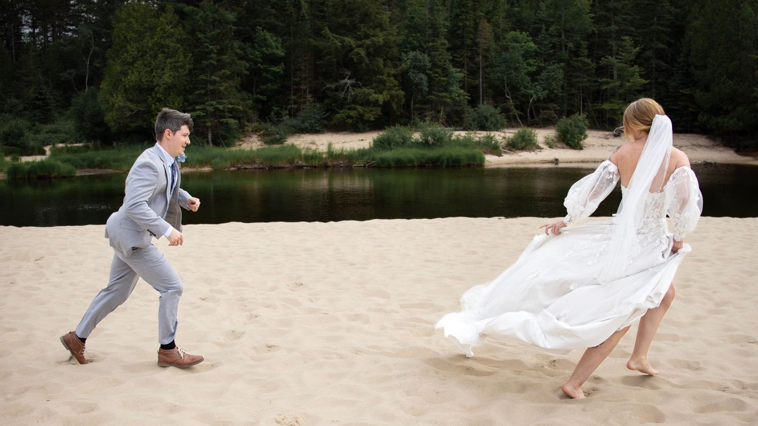 Bride and groom playfully running on sandy beach near forest lake, bride's white dress flowing in the wind.