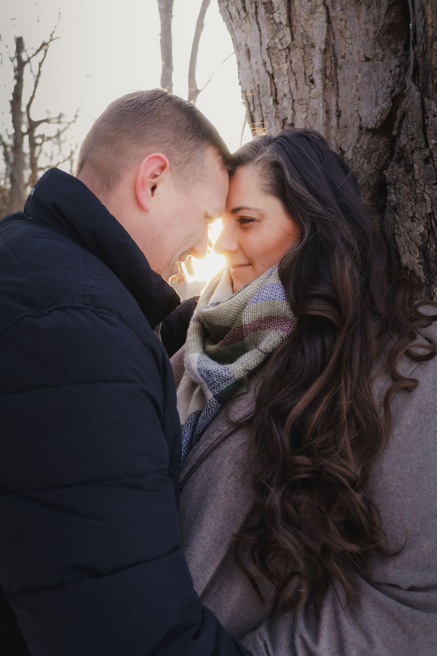 A couple standing close together beside a tree during an outdoor winter engagement session with soft sunlight.