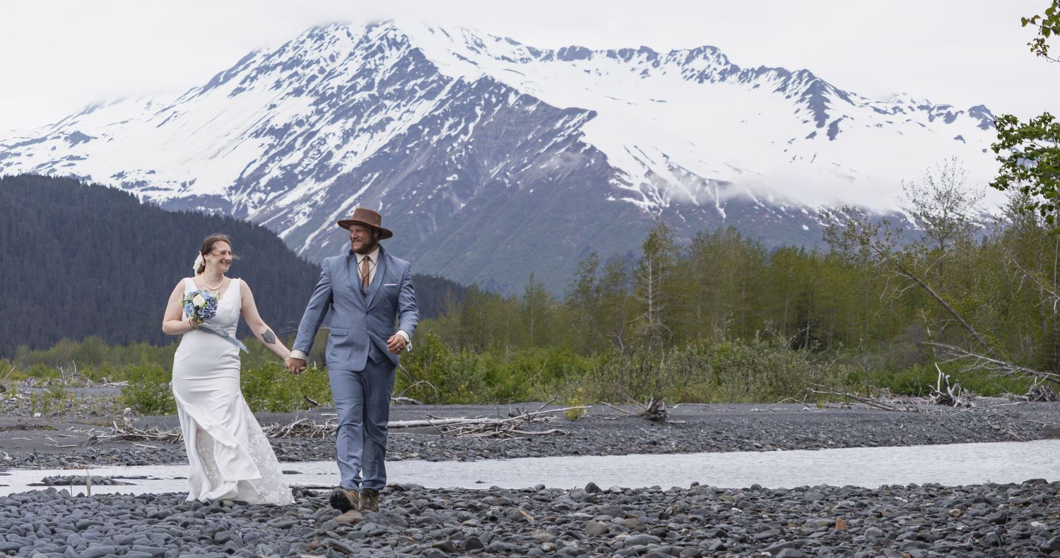 Couple in wedding attire walks hand-in-hand on rocky Alaskan terrain with snow-capped mountains in the background.