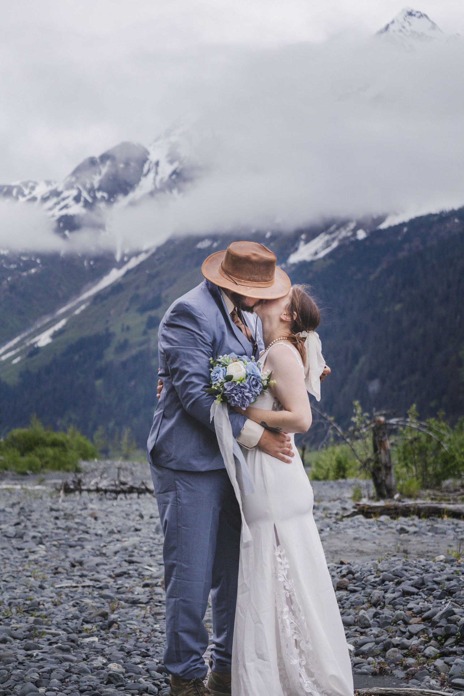 Bride and groom kissing in front of snow-capped mountains, groom wearing brown hat and blue suit, bride holding blue bouquet.