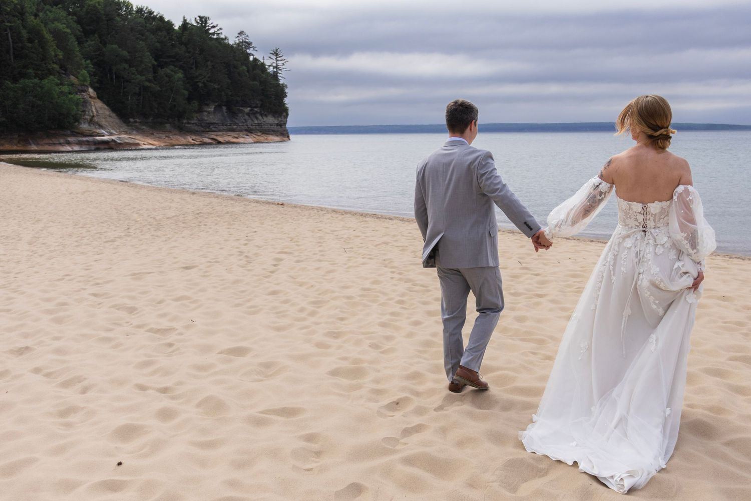 A bride and groom walk hand-in-hand along a sandy beach, with calm ocean waters and lush green trees in the background.